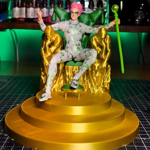 May include: A Riddler action figure sits on a golden throne with two golden statues. The figure wears a white suit with green question marks and has pink hair. A green staff with a question mark head is held in the figure's hand.