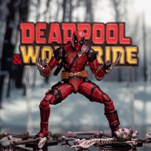 May include: A red and black action figure of Deadpool with a red and black suit, holding two silver claws. The figure is standing on a snowy surface with a sign that reads "Deadpool & Wolverine" in the background.