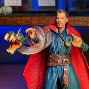 May include: A red and blue action figure of Doctor Strange with a translucent snake wrapped around his arm. The figure is wearing a red cape and a blue robe with a brown belt. The snake has a green head with sharp teeth.