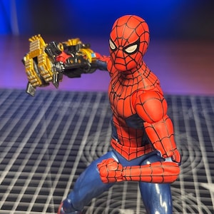 May include: A red and blue action figure of a character with a spider-web pattern on the red sections of the suit. The figure has white eyes and is posed with one arm extended forward, holding a metallic gold and black mechanical accessory.