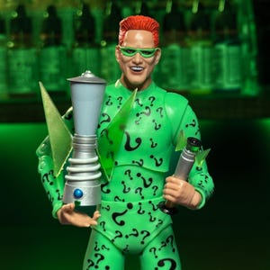 May include: A green action figure of the Riddler from Batman, wearing a green suit with question marks all over it. He is holding a silver and blue weapon in his left hand and a silver and blue weapon in his right hand.