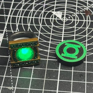 May include: A Green Lantern ring and a matching emblem. The ring is gold-colored with green accents and a glowing green center. The emblem is a green lantern symbol on a black circular base.