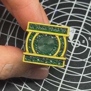 May include: A square-shaped ring featuring a central circular emblem with concentric rings, surrounded by a textured dark green surface and framed by a metallic gold border. The design includes horizontal gold bars at the top and bottom edges.