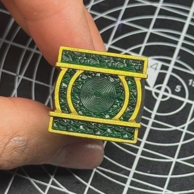 May include: A square-shaped ring featuring a central circular emblem with concentric rings, surrounded by a textured dark green surface and framed by a metallic gold border. The design includes horizontal gold bars at the top and bottom edges.