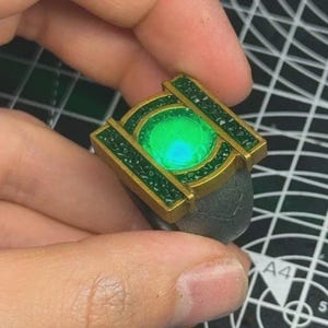 May include: A ring featuring a central circular glowing green light surrounded by a gold-colored frame with textured green accents. The band of the ring is dark gray with a cracked, weathered surface texture, creating a distinct contrast between the metallic gold, the bright green light, and the dark, rugged band.