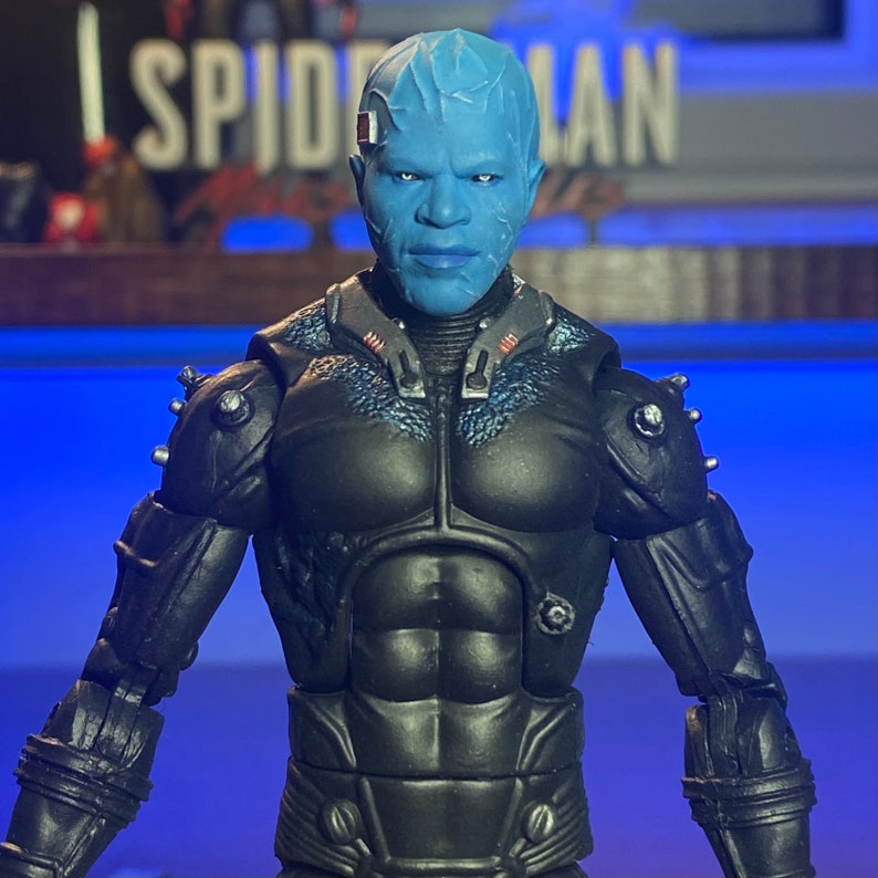 May include: A blue and black action figure of a superhero with a textured blue face and a black suit with silver accents. The figure is posed with its arms outstretched and its head tilted slightly to the side.