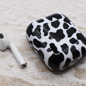 Op de afbeelding: Een witte en zwarte koe-print telefoonhoesje voor AirPods. De hoes is gemaakt van hard plastic en heeft een glanzende afwerking. De hoes wordt getoond naast een witte AirPod.