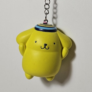 May include: A yellow keychain featuring a cartoon character with a black smiling face, small arms, and a blue and white striped hat. The keychain is attached to a silver metal ring and chain.