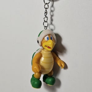 May include: A keychain featuring a cartoon character, a turtle-like creature with a yellow body, green shell, and green shoes. The character wears a white helmet with a green stripe. The keychain has a silver chain and ring.