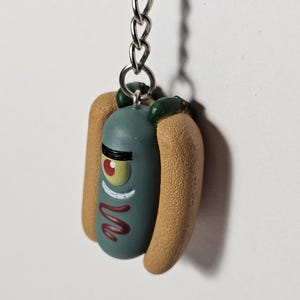 Op de afbeelding: Een novelty sleutelhanger met een stripfiguur in de vorm van een hotdog. Het personage is teal met één oog, een rode mond en een zwarte wenkbrauw. De hotdogbroodjes zijn bruin en de sleutelhanger heeft een zilveren ketting.