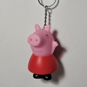 May include: A Peppa Pig keyring featuring a pink pig in a red dress and black shoes. The keyring is attached to a silver chain and ring, ready for keys or a bag.