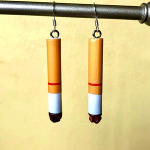 Op de afbeelding: Een paar novelty oorbellen die op sigaretten lijken. Elke oorbel heeft een cilindrische vorm met een bruin lichaam, een witte punt, een rode band en een donkerrode, getextureerde punt. De oorbellen hangen aan zilverkleurige haken.