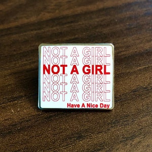 May include: A white enamel pin with a red outline and the text "NOT A GIRL" repeated multiple times. The pin also has the text "Have A Nice Day" at the bottom.
