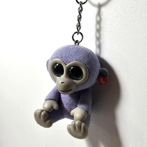 May include: A lilac monkey keyring with big black eyes, grey hands and feet, and a red heart-shaped tag. The plush toy is attached to a silver chain and keyring.