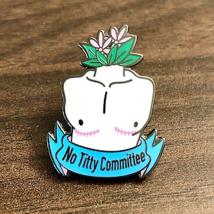 Op de afbeelding: Een witte emaille pin met een gestileerde torso met een bloemenkrans en een banner met de tekst "No Titty Committee". De pin heeft een zwarte omtrek en roze accenten, inclusief de bloemen en de tekst op de banner.