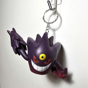 May include: A purple and red keychain featuring a cartoon character with a single yellow eye, a wide grin, and spiky features. The keychain is attached to a silver keyring and hangs from a clear adhesive hook.