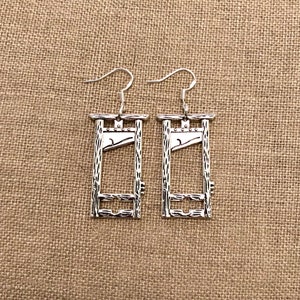 Puede incluir: Un par de pendientes de plata en forma de guillotina. Los pendientes tienen forma de guillotina con una cuchilla y un marco de madera. Los pendientes cuelgan de ganchos de plata.