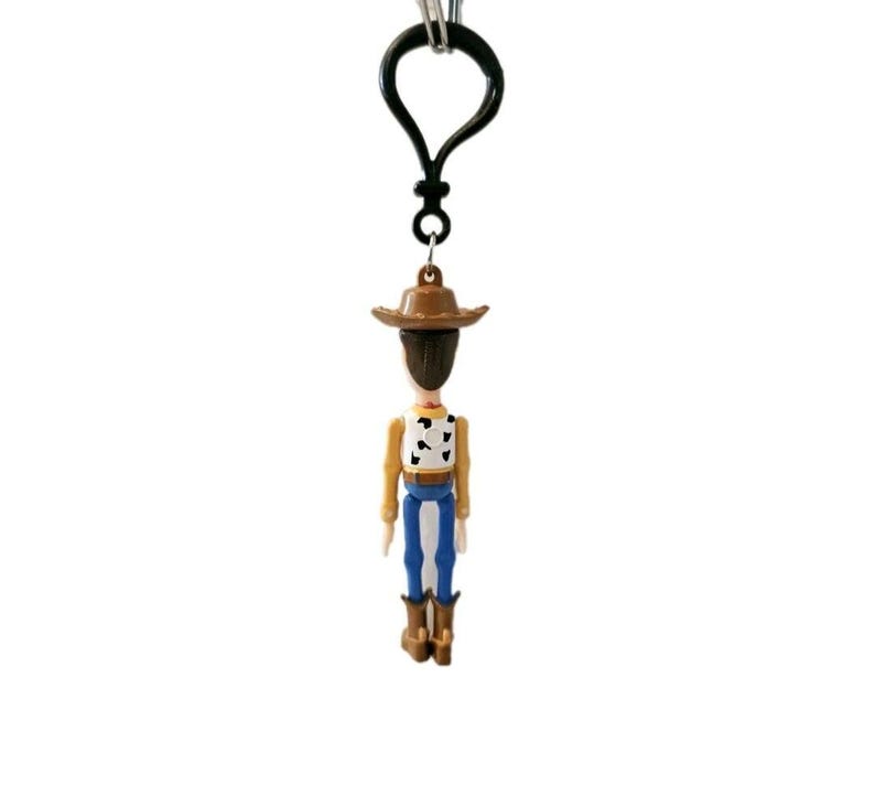 May include: A Woody character keychain. The miniature figure has a brown cowboy hat, a tan shirt with a black and white cow pattern, blue pants, and brown boots. The keychain has a black clasp.