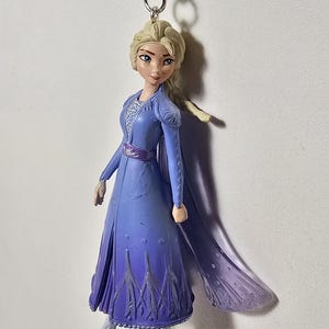 May include: A figurine of a character from the animated film "Frozen", featuring a blonde woman in a long blue dress with a purple ombre effect. The figure has a silver chain for hanging and is wearing silver boots.