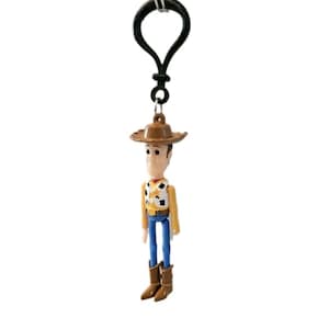 May include: A Woody keychain featuring a miniature figurine of the character from the animated film. The figure wears a brown cowboy hat, a yellow vest with a black and white cow pattern, blue pants, and brown boots. The keychain has a black metal clasp.
