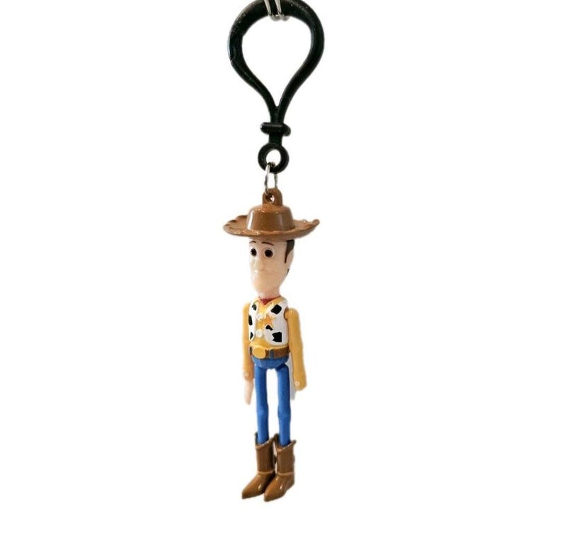 May include: A Woody keychain featuring a miniature figurine of the character from the animated film. The figure wears a brown cowboy hat, a yellow vest with a black and white cow pattern, blue pants, and brown boots. The keychain has a black metal clasp.