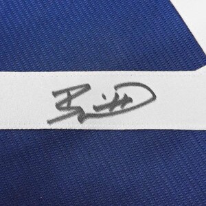 May include: Close-up of a blue fabric with a white stripe. A signature is visible on the white stripe. The fabric has a textured appearance. The signature is in black ink.