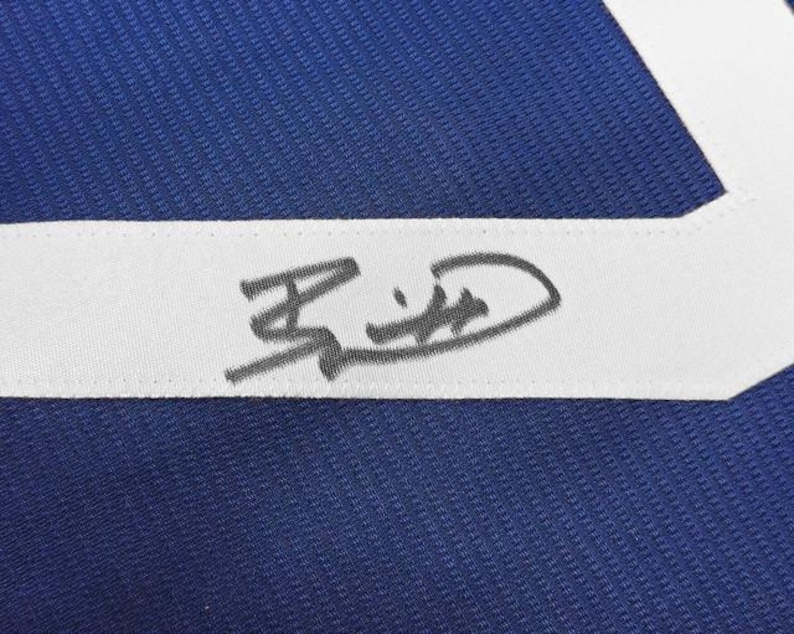 May include: Close-up of a blue fabric with a white stripe. A signature is visible on the white stripe. The fabric has a textured appearance. The signature is in black ink.