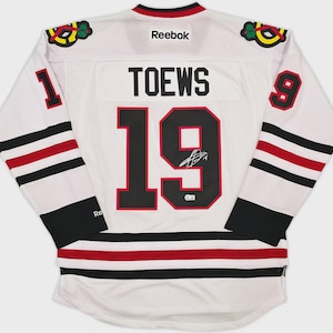 May include: A white Reebok hockey jersey featuring the name TOEWS and the number 19 in black with red outlines on the back. The sleeves display the number 19, black and red stripes, and team logos. A signature is visible on the number 19.