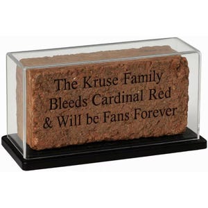May include: A rectangular red clay brick encased in a clear acrylic display box with a black base. The brick features the engraved text: The Kruse Family Bleeds Cardinal Red & Will be Fans Forever. The display case has a transparent top and sides.