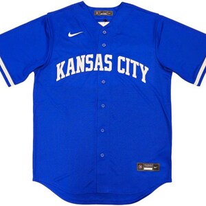May include: A royal blue baseball jersey with the words "KANSAS CITY" in white across the chest. The jersey has a button-up front, white stripes on the sleeves, and a small Nike logo. The jersey is a size 46.