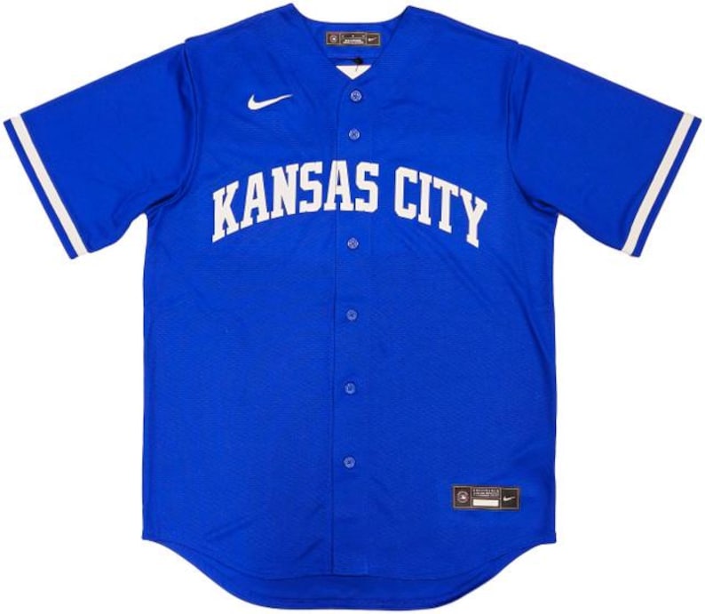 May include: A royal blue baseball jersey with the words "KANSAS CITY" in white across the chest. The jersey has a button-up front, white stripes on the sleeves, and a small Nike logo. The jersey is a size 46.