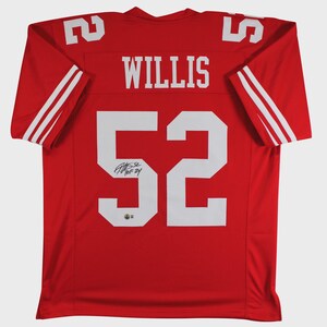 May include: A red sports jersey featuring the name WILLIS and the number 52 in large white block lettering on the back. The sleeves have white stripes, and the number 52 includes a black ink signature and the text HOF 24 in the lower section.