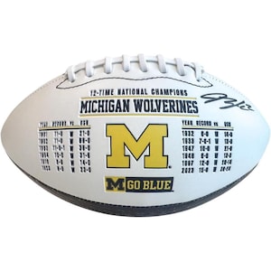 May include: A white American football featuring the text 12-TIME NATIONAL CHAMPIONS MICHIGAN WOLVERINES, a yellow block M logo, the text GO BLUE, and a list of historical records against OSU, along with a black signature on the right side.