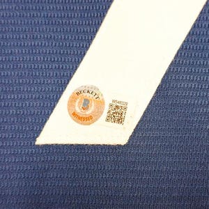 May include: Close-up of a blue textured fabric with a white angled stripe. A Beckett Witnessed sticker is affixed to the white stripe, along with a QR code and the number W548329.
