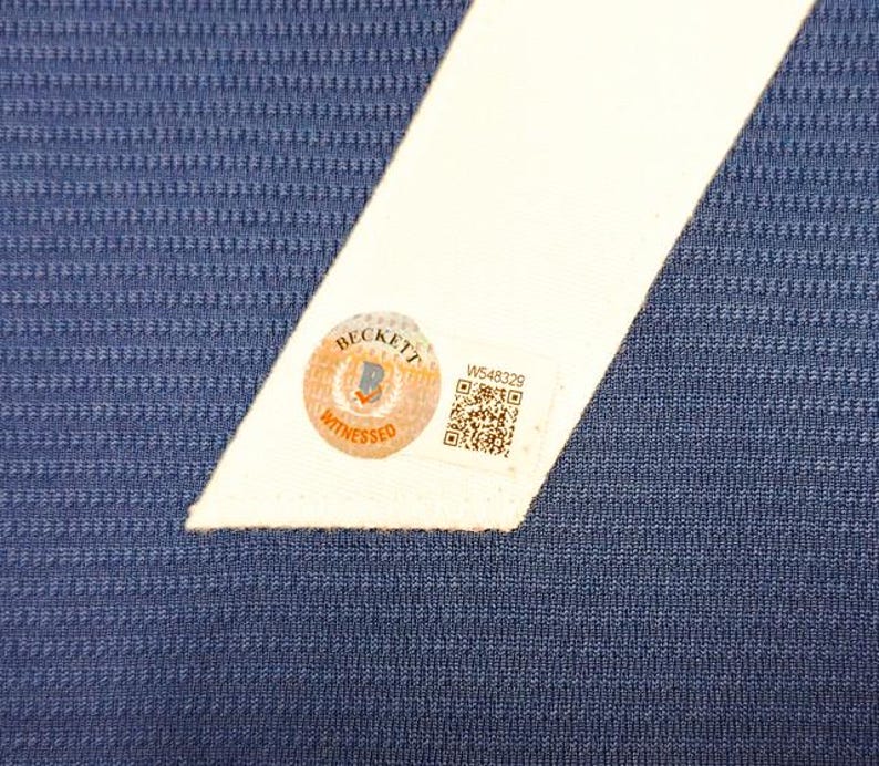 May include: Close-up of a blue textured fabric with a white angled stripe. A Beckett Witnessed sticker is affixed to the white stripe, along with a QR code and the number W548329.