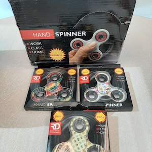 May include: Three packages of hand spinners. The spinners are made of plastic and have different designs. The packages are black with white text and images. The text on the packages says "Hand Spinner" and "Emoji".