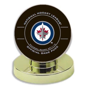 May include: A gold-colored hockey puck holder with a black and brown puck. The puck features the text "National Hockey League" and "Official Game Puck". The center of the puck has a logo with a maple leaf.