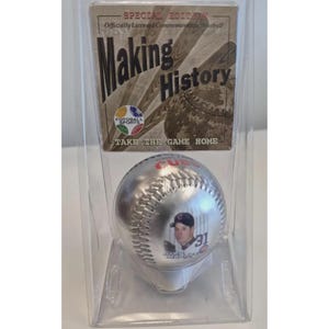 May include: A silver commemorative baseball featuring a player's image and the number 31. The baseball is in a clear plastic case with a cardboard header that reads "Making History" and "Take the Game Home."