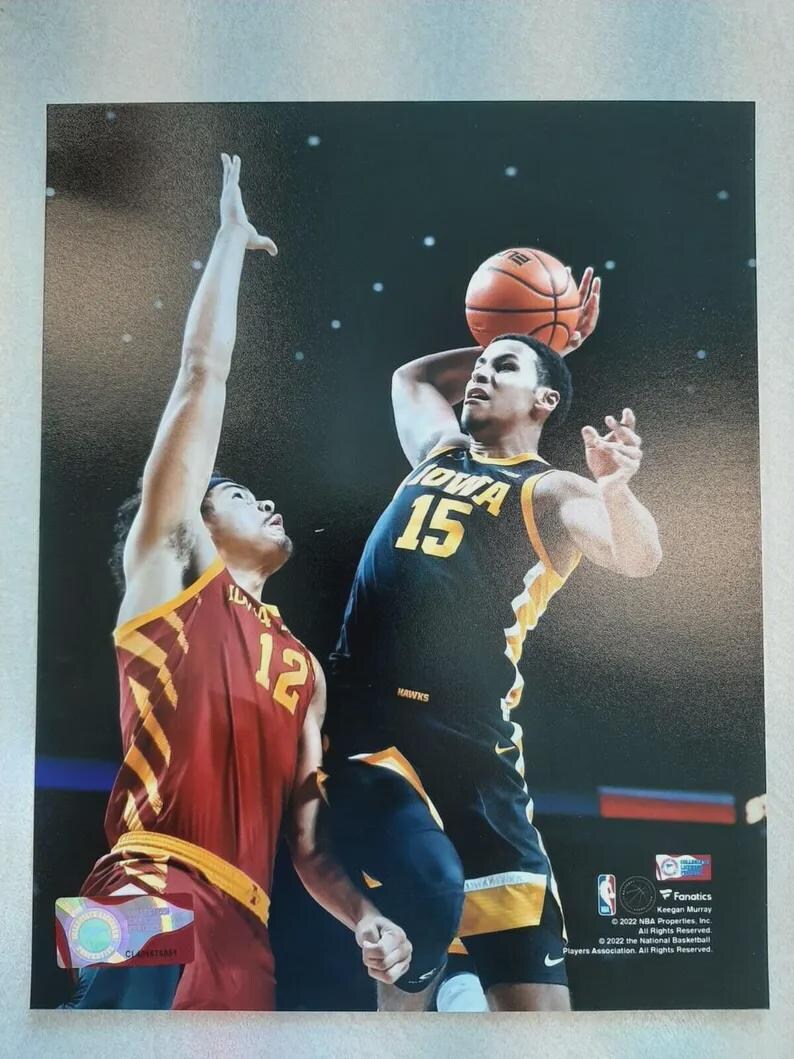 Puede incluir: Imagen de acci&oacute;n de baloncesto con dos jugadores en el aire. Un jugador, con una camiseta negra y amarilla con el n&uacute;mero 15, est&aacute; a punto de lanzar el bal&oacute;n. El otro jugador, con una camiseta roja y dorada con el n&uacute;mero 12, intenta bloquear el tiro.
