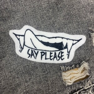 May include: White embroidered patch with a black outline of a mouth with fangs and a tongue sticking out. The text "Say Please" is written below the mouth.