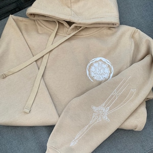 May include: A tan hoodie with a white embroidered design on the front and a white embroidered sword design on the sleeve.