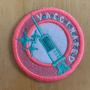 May include: A pink and white embroidered patch with the text "Vaccinated" in a circle around a turquoise syringe with three stars.