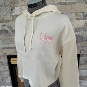 May include: A cream-colored cropped hoodie with a pink heart and the word "Anthony" embroidered on the front.