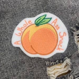May include: Embroidered patch featuring a peach with the text "A Whole Ass Snack". The peach is in shades of orange and yellow with green leaves. The patch is on a white background and set against a denim background.