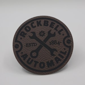 May include: A brown leather patch with black text and a graphic of two crossed wrenches. The text reads "Rockbell Automail - Est'd 1884 - "