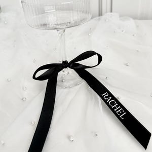 May include: A clear champagne coupe with a ribbed bowl and slender stem. A black ribbon is tied around the stem, with the name "RACHEL" printed in white. The glass sits on a white, pearl-studded fabric.