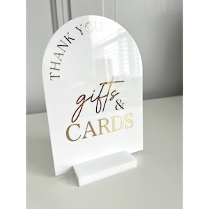 May include: A white acrylic sign with gold lettering that reads "THANK YOU gifts & CARDS". The sign has an arched top and a rectangular base for standing. The text is in a stylish script font.