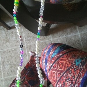 Może przedstawiać: Biały naszyjnik z koralików z kolorowymi koralikami i koralikami z literami, które tworzą napis "LOVE", wiszący na krześle. Naszyjnik leży na wzorzystym tle z tkaniny.