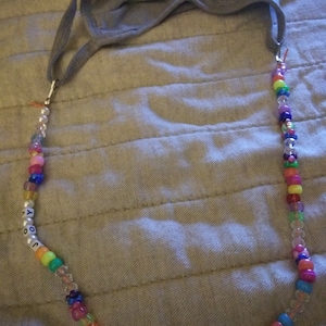 Może przedstawiać: Kolorowy sznurek z koralikami z imieniem "JOOKY" napisanym białymi koralikami. Sznurek jest przymocowany do szarej maseczki z tkaniny.