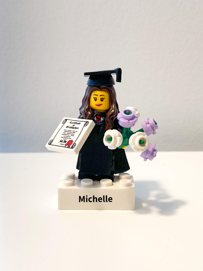 May include: A Lego figurine of a graduate wearing a black cap and gown, holding a diploma and a bouquet of white and purple flowers. The base of the figurine is white with the name 'Michelle' printed on it.